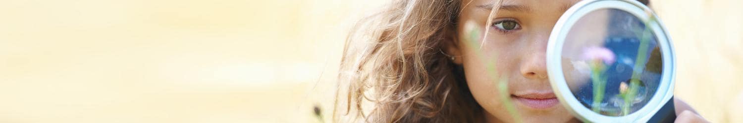 Ein Mädchen mit langen Haaren hält eine Lupe vor ihr Gesicht und fokussiert auf ein kleines Objekt vor hellem Hintergrund.