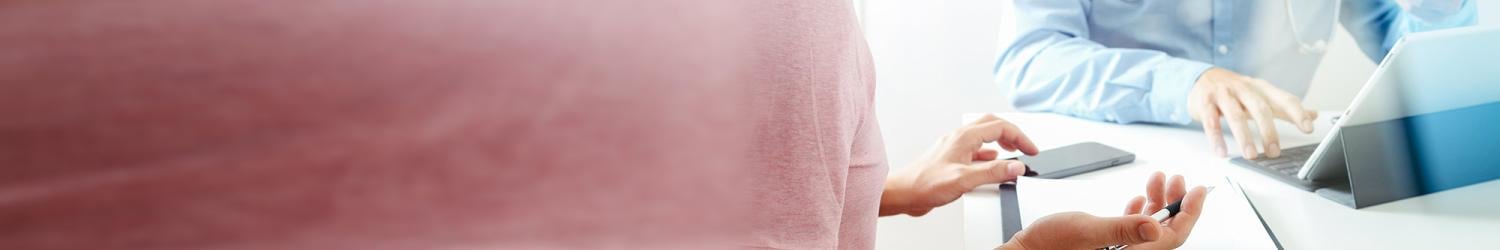 Eine Person im rosa Hemd konsultiert einen Arzt im blauen Hemd, benutzt einen Laptop und hält einen Stift.