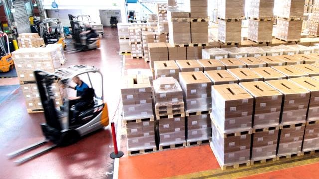 Belebtes Lager mit Stapeln von Kartons und einem Arbeiter, der einen Gabelstapler bedient.