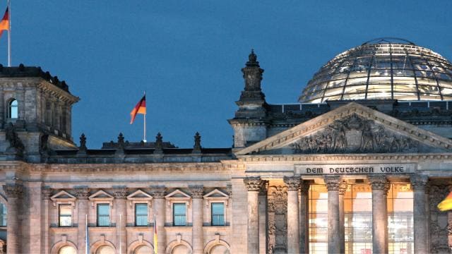 Das beleuchtete Reichstagsgebäude in Berlin in der Abenddämmerung mit deutlich sichtbaren deutschen Flaggen.
