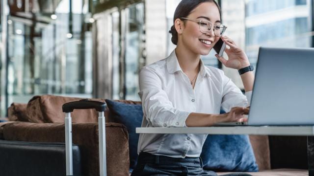 Eine Frau im weißen Hemd benutzt in einer Flughafenlounge einen Laptop und telefoniert mit einem Smartphone. Neben ihr steht ein Koffer.