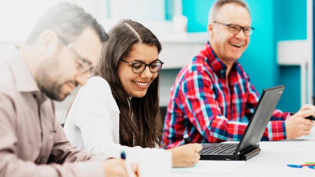 Drei Personen sitzen an einem Tisch. Die Person links schreibt, die Person in der Mitte lächelt, während sie einen Laptop benutzt, und die Person rechts lächelt, trägt eine Brille und ein rotkariertes Hemd.