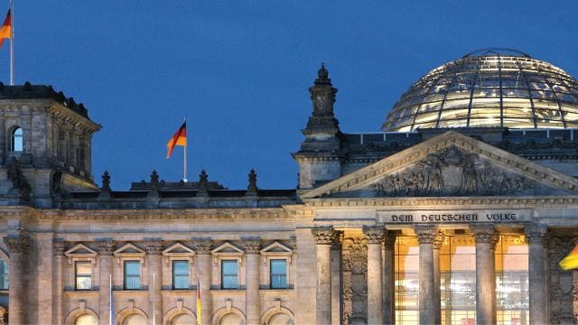 Das Reichstagsgebäude in Berlin in der Abenddämmerung mit einer Glaskuppel und mehreren deutschen Flaggen.
