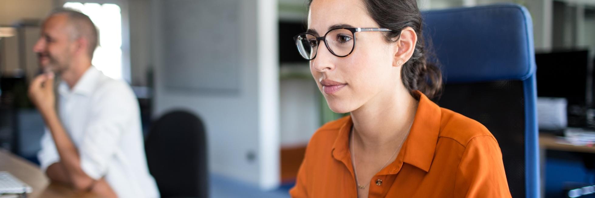 Eine Frau mit Brille und orangefarbenem Hemd arbeitet in einem modernen Büro an einem Computer, während im Hintergrund ein Mann sitzt.