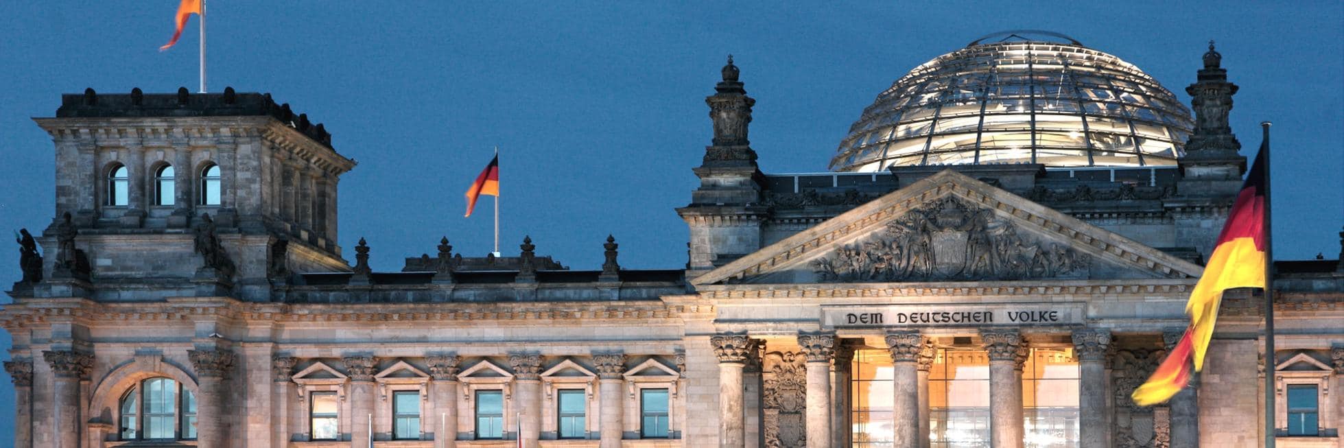 Das beleuchtete Reichstagsgebäude in Berlin in der Abenddämmerung mit deutlich sichtbaren deutschen Flaggen.