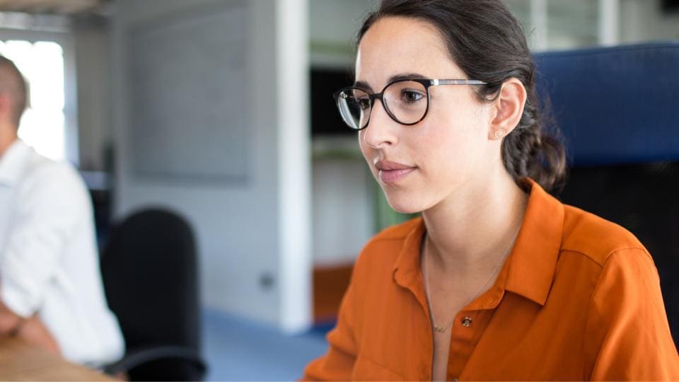 Eine Frau mit Brille und orangefarbenem Hemd arbeitet in einem modernen Büro an einem Computer, während im Hintergrund ein Mann sitzt.