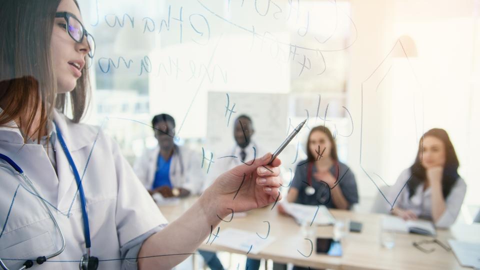 Eine Person im weißen Kittel schreibt Diagramme auf eine Glastafel, während im Hintergrund eine Gruppe von Menschen an einem Tisch sitzt.
