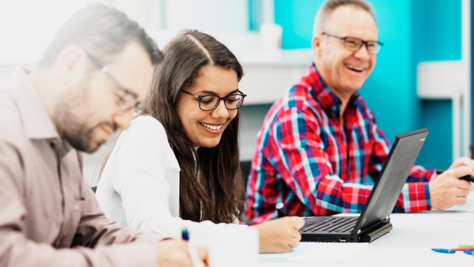 Drei Personen sitzen an einem Tisch. Die Person links schreibt, die Person in der Mitte lächelt, während sie einen Laptop benutzt, und die Person rechts lächelt, trägt eine Brille und ein rotkariertes Hemd.