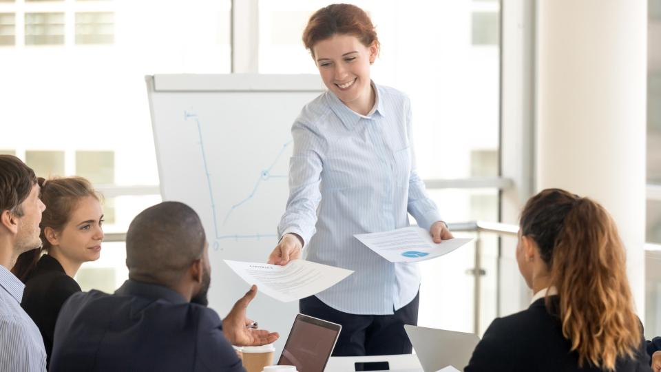 Eine Frau verteilt in einem Besprechungsraum Unterlagen an Kollegen. Im Hintergrund ist ein Flipchart mit einer Grafik zu sehen.