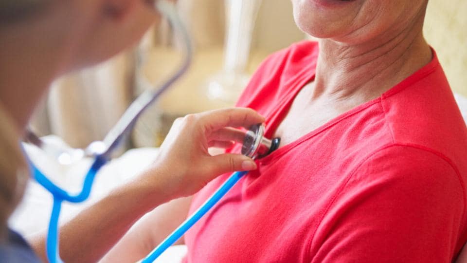Eine medizinische Fachkraft untersucht mit einem Stethoskop das Herz einer lächelnden Patientin, die ein Kopftuch und ein rotes Hemd trägt.