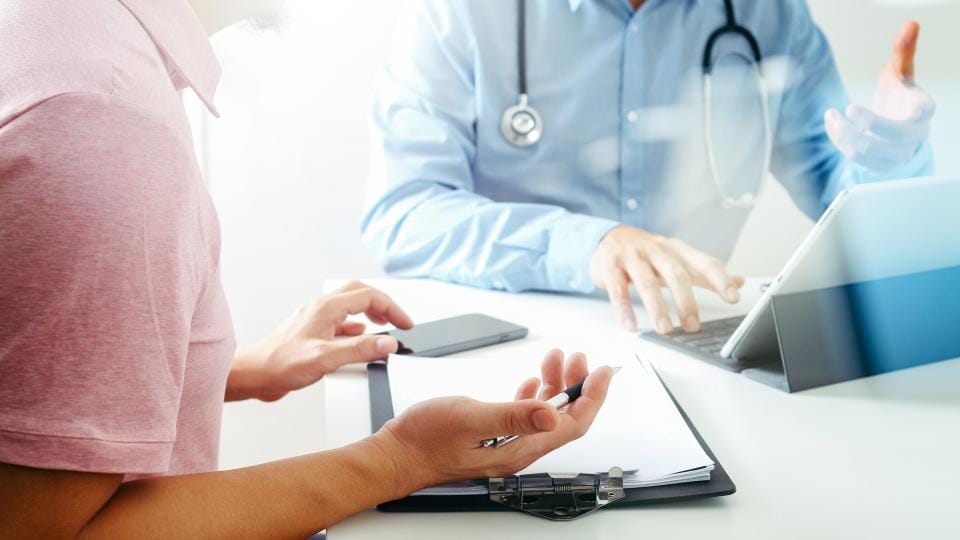 Ein Arzt und ein Patient sitzen während einer Konsultation mit einem Tablet, einem Smartphone und einem Klemmbrett an einem Schreibtisch.