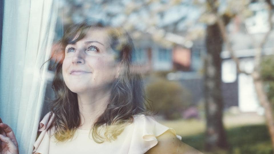 Eine Frau mit langen Haaren steht an einem Fenster und schaut nach oben. Die Spiegelung zeigt eine verschwommene Außenszene mit Bäumen und einem Gebäude.