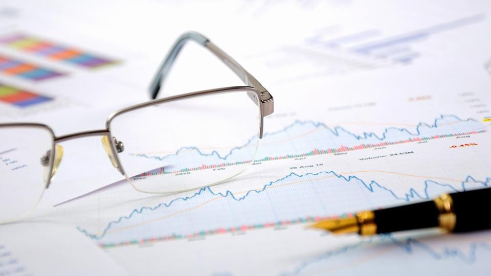 Eine Brille und ein Stift liegen auf Finanzdiagrammen mit Liniendiagrammen und Daten im Hintergrund.