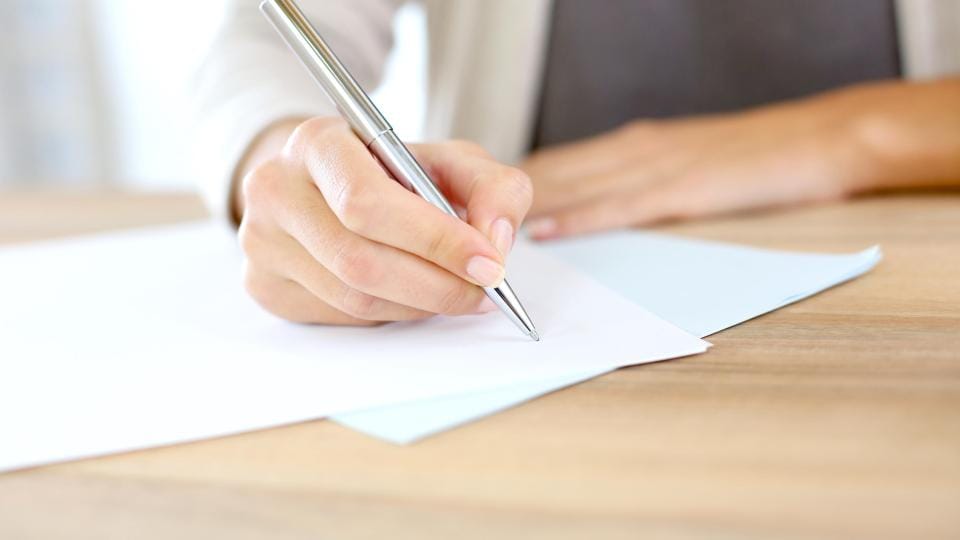 Eine Person schreibt mit einem silbernen Stift auf ein Blatt Papier, das auf einem Holztisch liegt.
