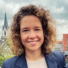 Person mit lockigem Haar, die im Freien lächelt und einen Blazer trägt. Im Hintergrund sind ein Kirchturm und Gebäude zu sehen.